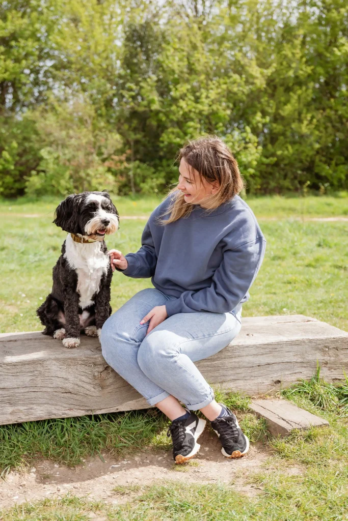 A woman in a blue jumper and light jeans sits on a wooden log bench in a grassy park, smiling down at a small black and white Tibetan Terrier sitting beside her. Leafy green trees fill the background on a sunny day.